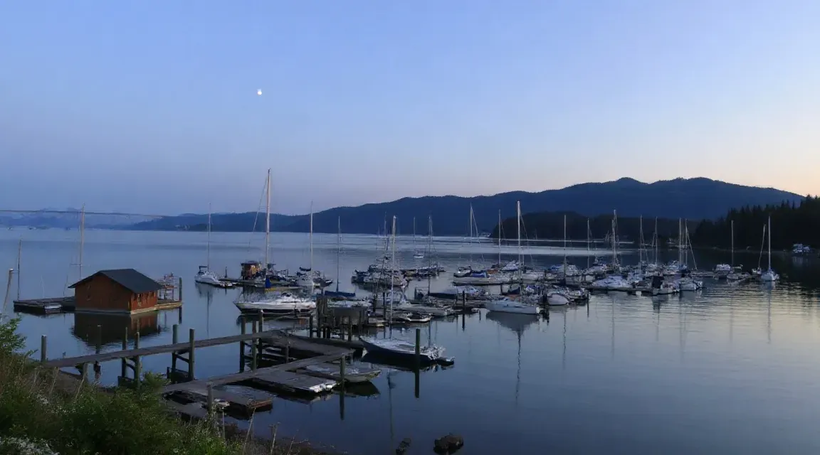 夕暮れの湖面に浮かぶ小さな船小屋と、遠くに並ぶ帆船